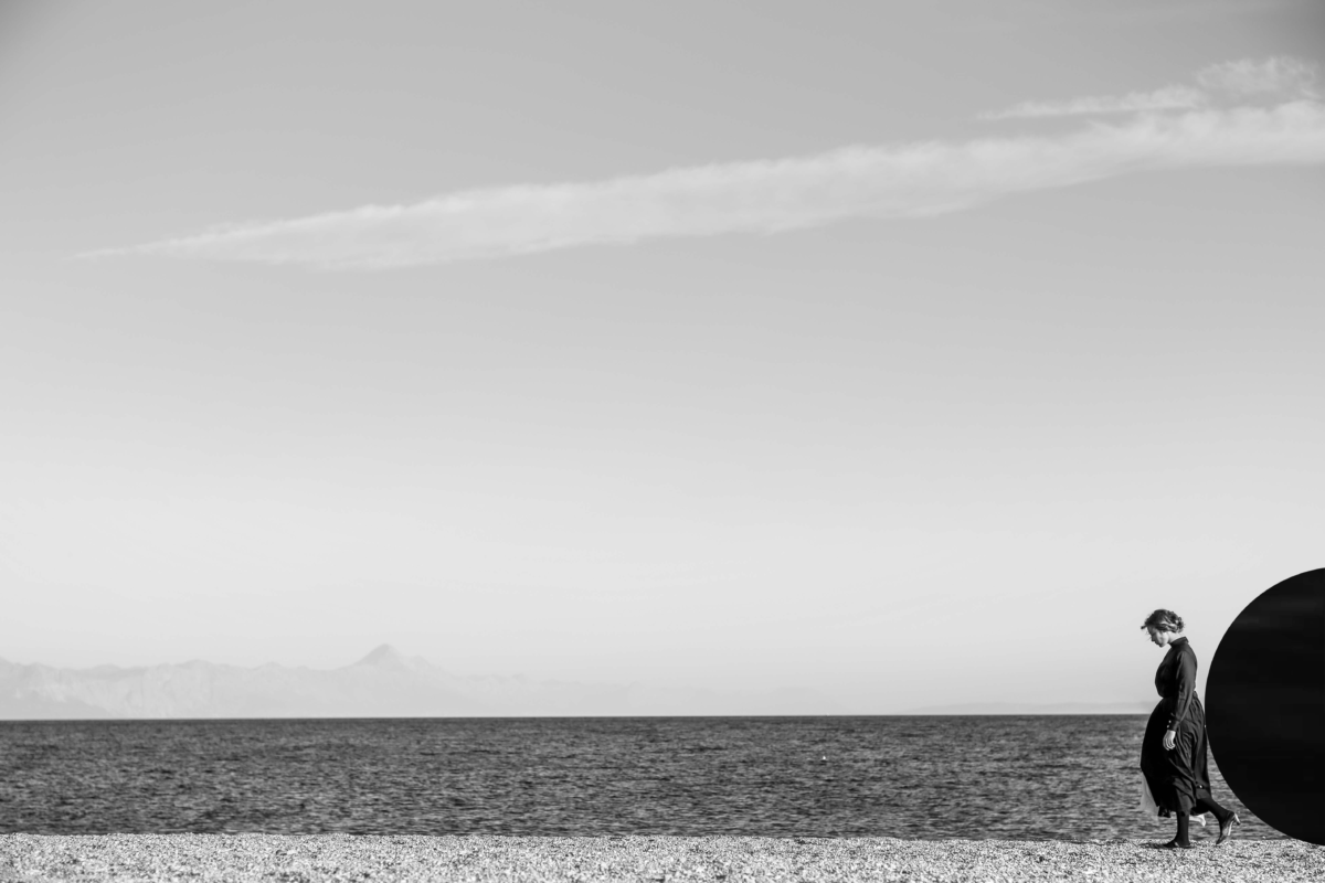 Schwarzweißfoto von einer Person vor dem Horizont des Meeres mit einem schwarzen, runden Objekt auf dem Rücken