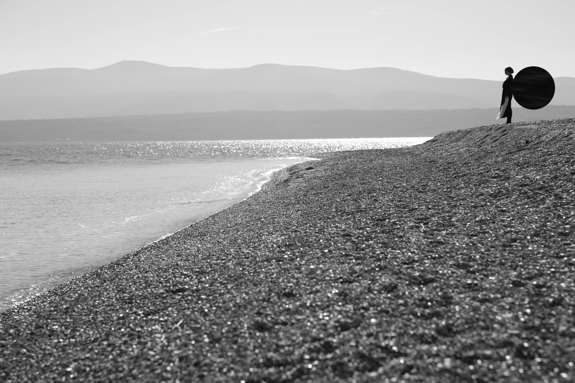 Schwarzweißfoto von einer Person an einem Kiesstrand mit einem schwarzen, runden Objekt auf dem Rücken