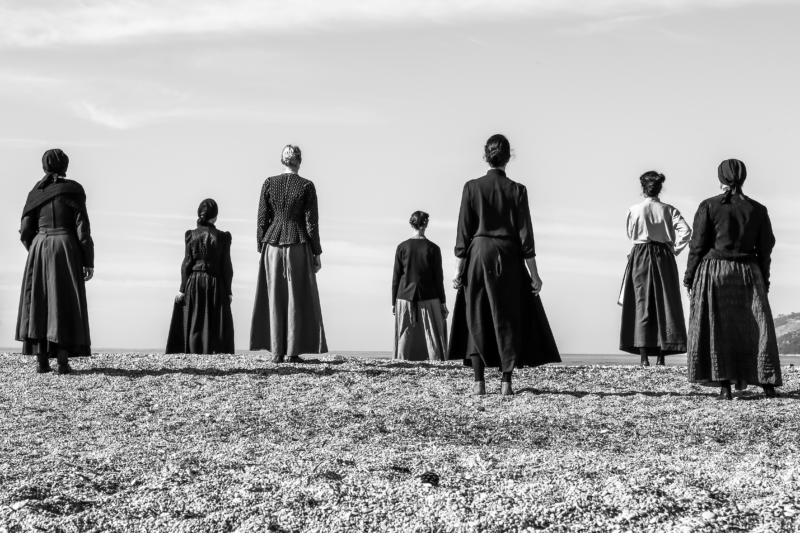 Filmstill in schwarzweiß von sieben stehenden weiblich gelesende Personen von hinten, die auf das Meer hinausblicken.