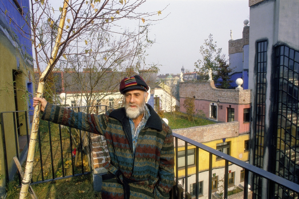 Ein Mann mit weißem Vollbart und Kappe auf einer Terrasse mit einem Baum in der Hand, im Hintergrund eine bunte Fassade