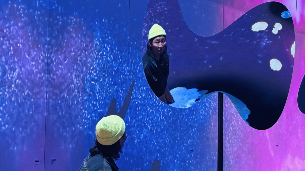 Künstlerin blickt in einen organisch geformten Spiegel vor mehrfarbigem blau-violettem Hintergrund