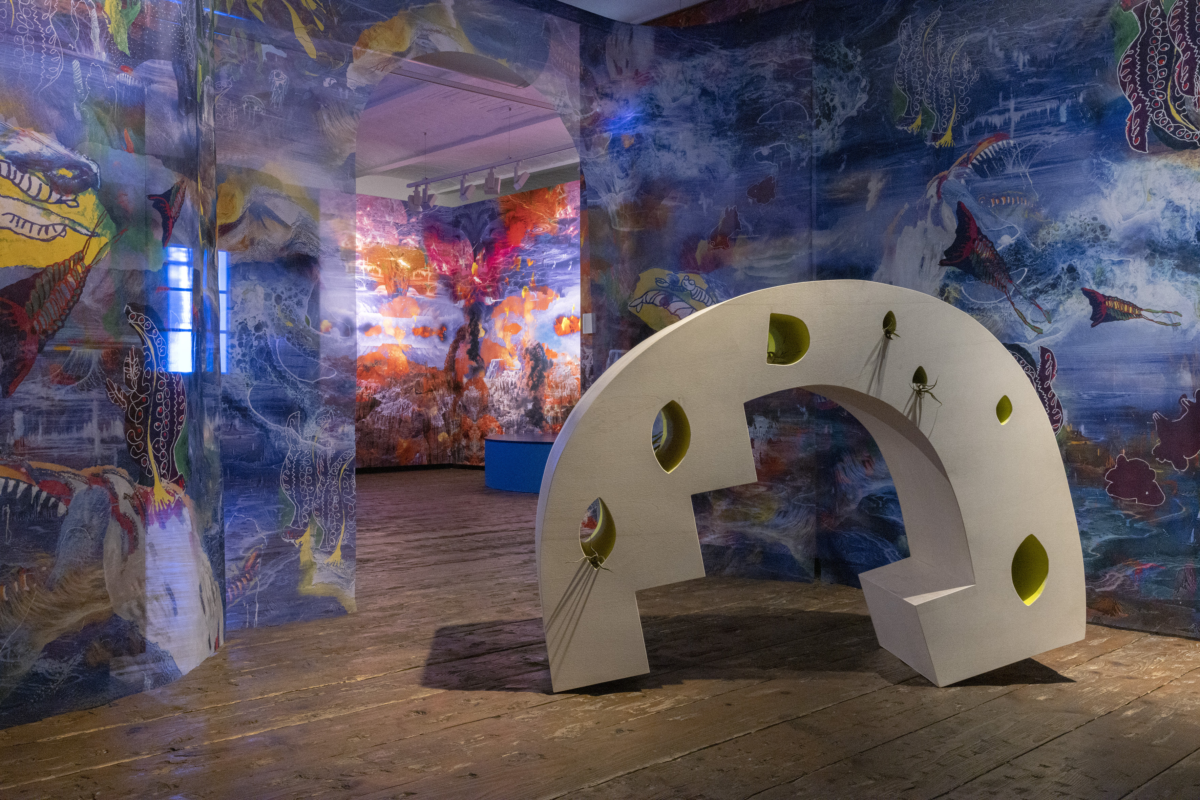 Halbkreisförmige, cremefarbene Skulptur mit rechteckigen Eckteilen auf einem Holzboden, umgeben von dünnen, durchsichtigen, mehrfarbigen Vorhängen