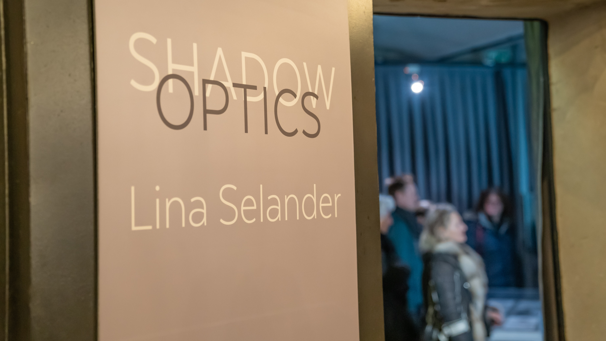 Ein Schild verkündet „Shadow Optics Lina Selander“, im Hintergrund sind Personen verschwommen zu sehen.
