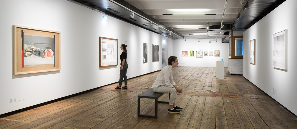 Zwei Personen, eine sitzend und eine stehend, betrachten Bilder im Ausstellungsraum