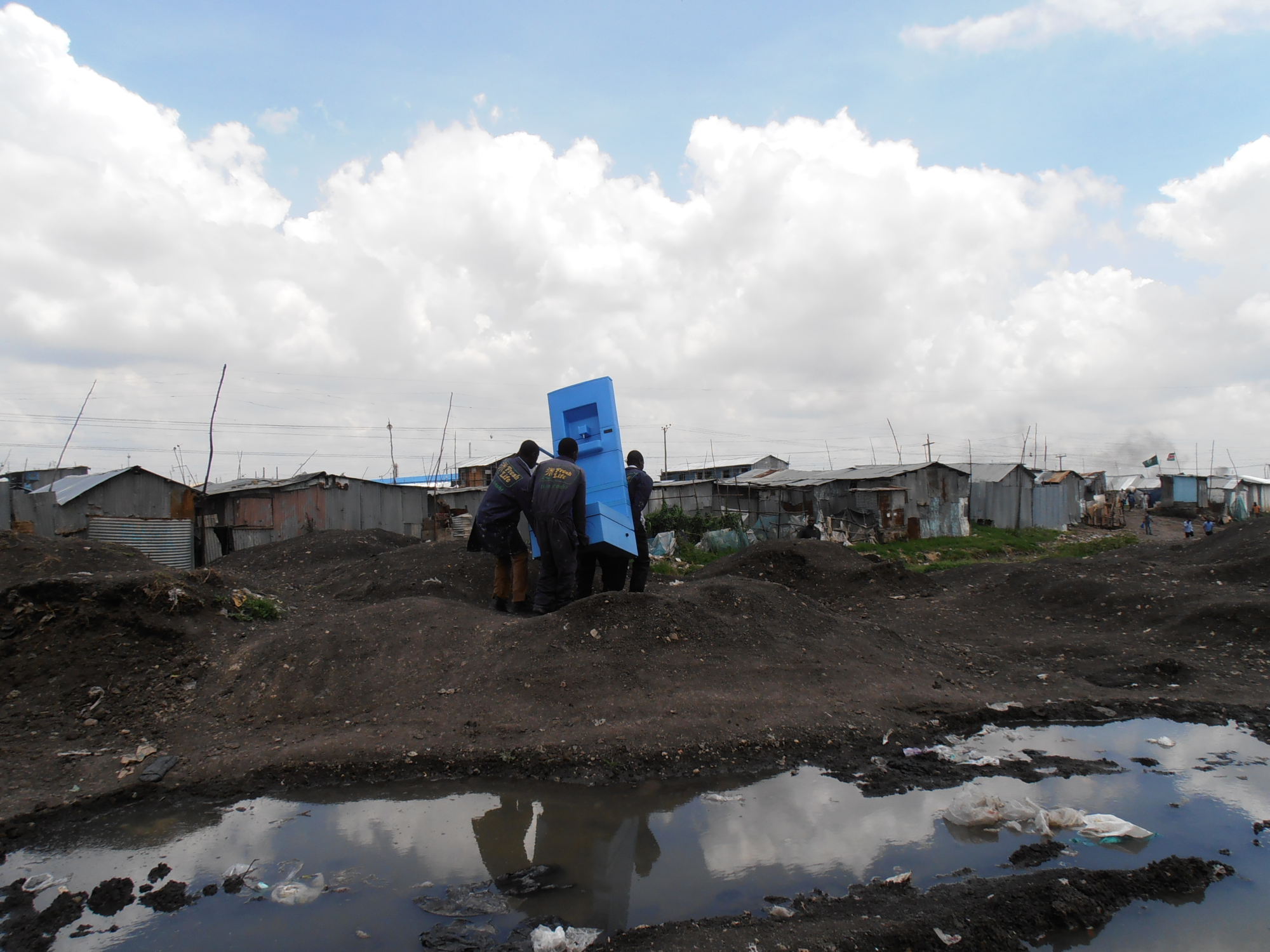 Three people carrying a blue object in a rural area next to a big, dirty puddle